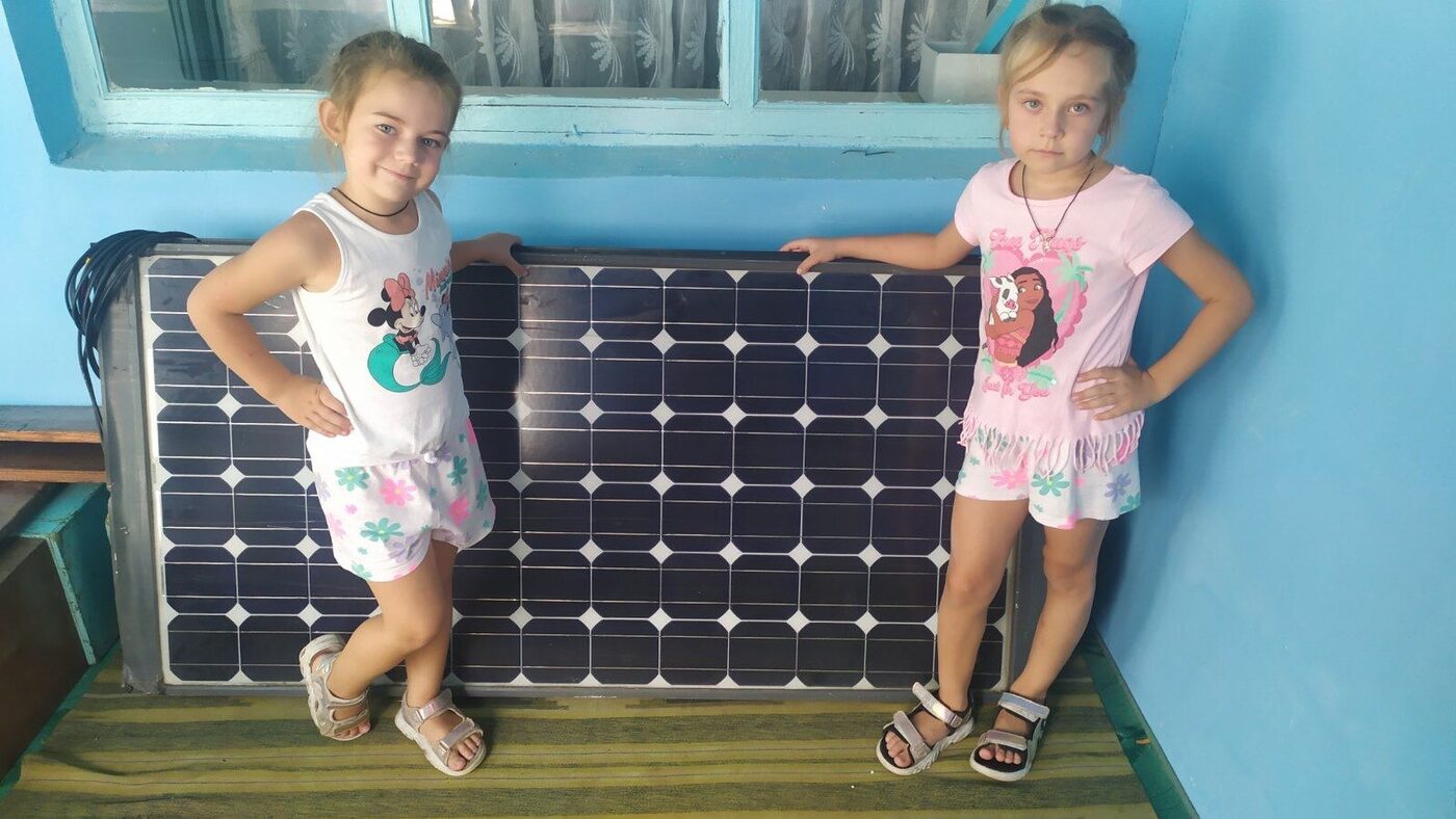 Two girls stand on each side of a solar panel leaning against a blue wall