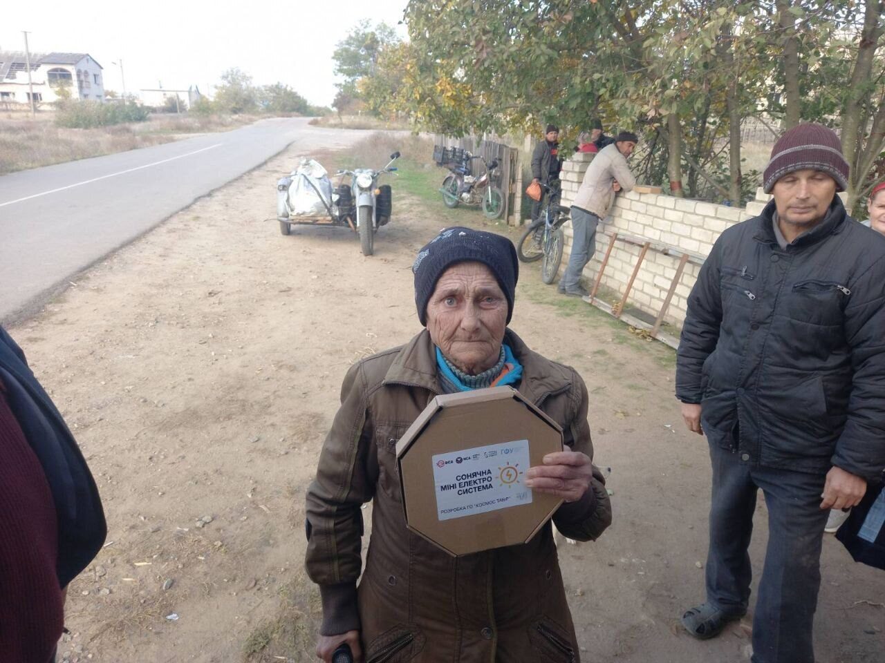 An elderly woman holds a MINISOLAR box on a village road
