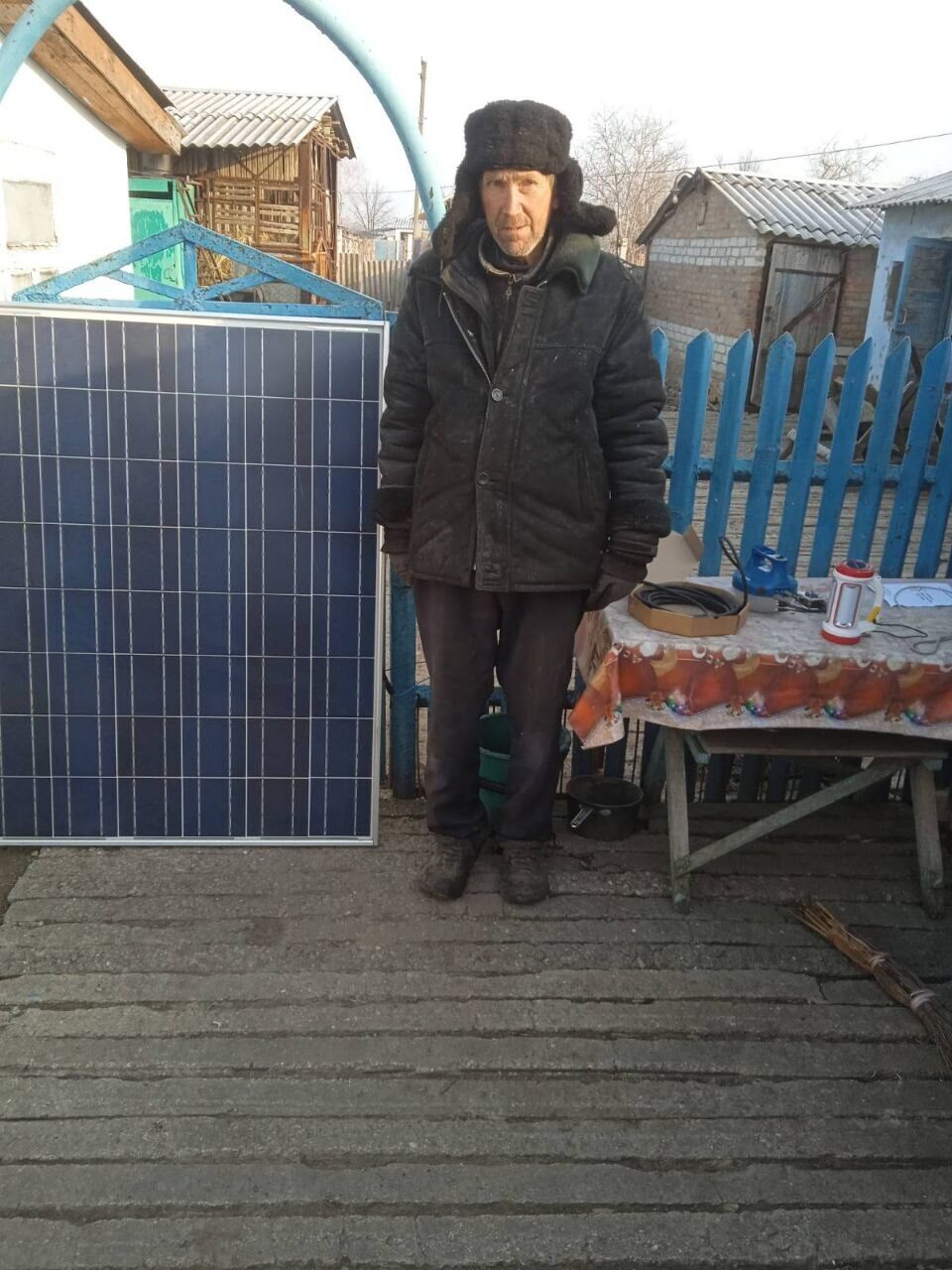 A man in a fur hat stands next to a solar panel on a porch